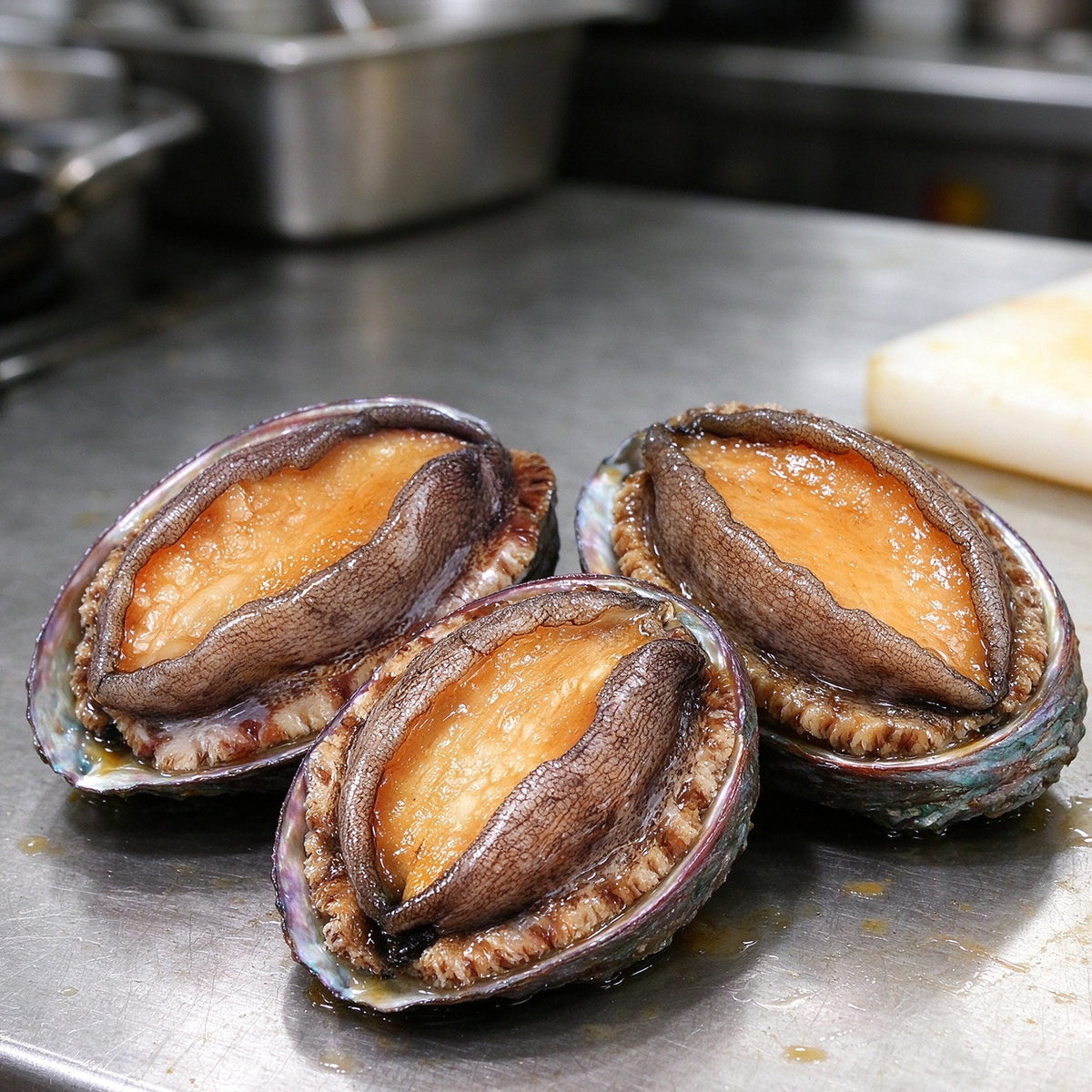 Whole abalone in dark shells with golden foot meat, resting on a steel bench